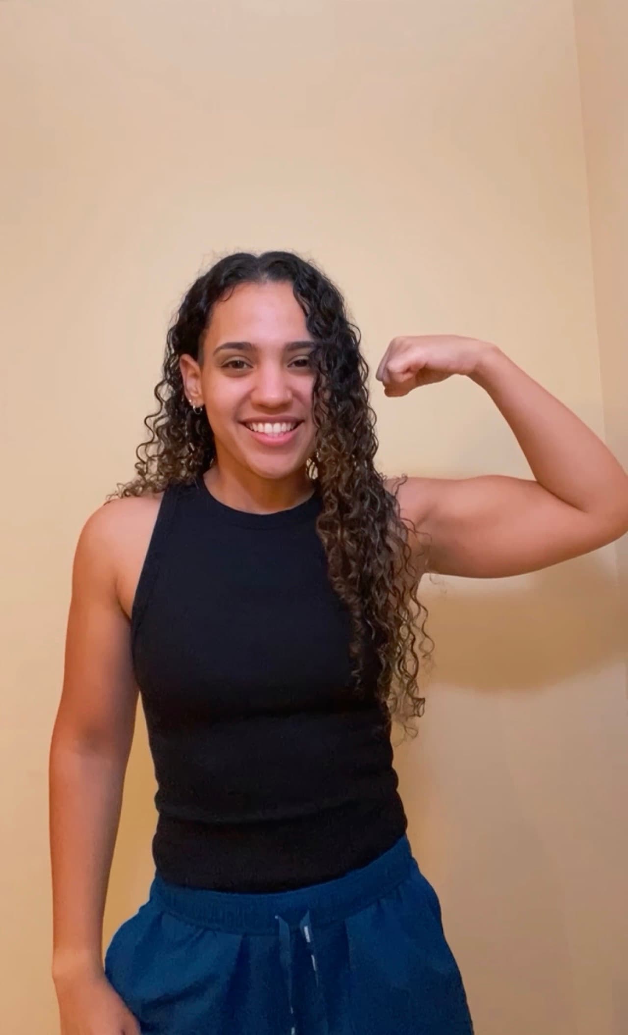 Smiling woman with long curly hair flexing her bicep in a black tank top.